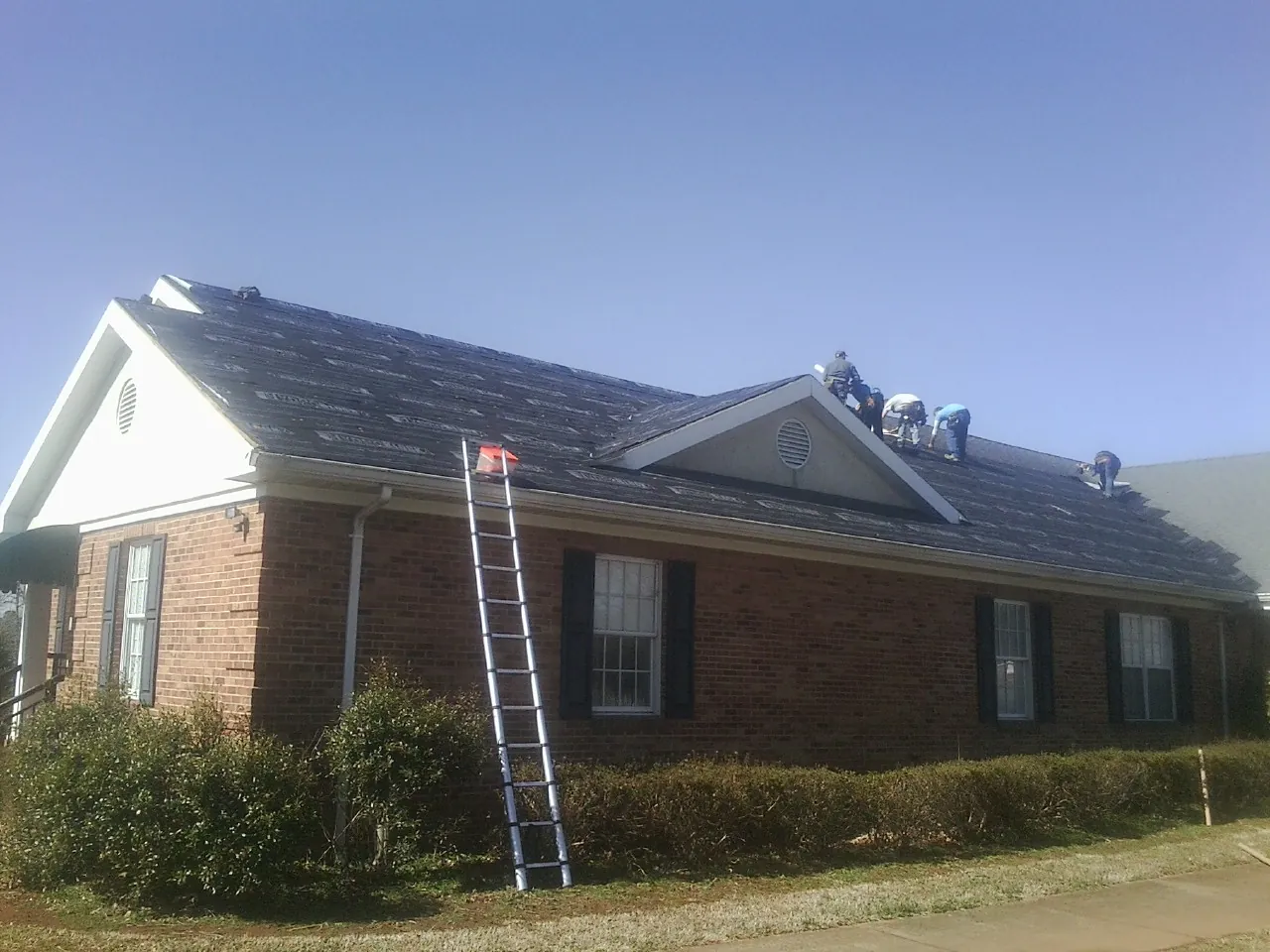 Crew tearing shingles off a brick ranch roof with a ladder set — Metal Roof Repair work by our crew in Yoakum