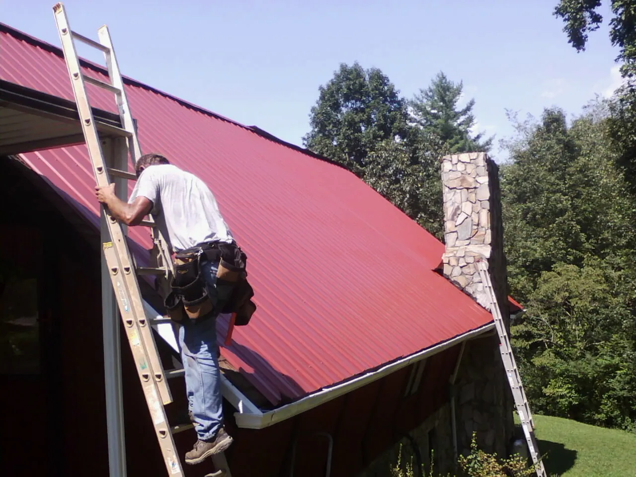 Roofer on a ladder beside a red metal roof and stone chimney — Soffit & Fascia Repair by Roofing Brothers in Yoakum